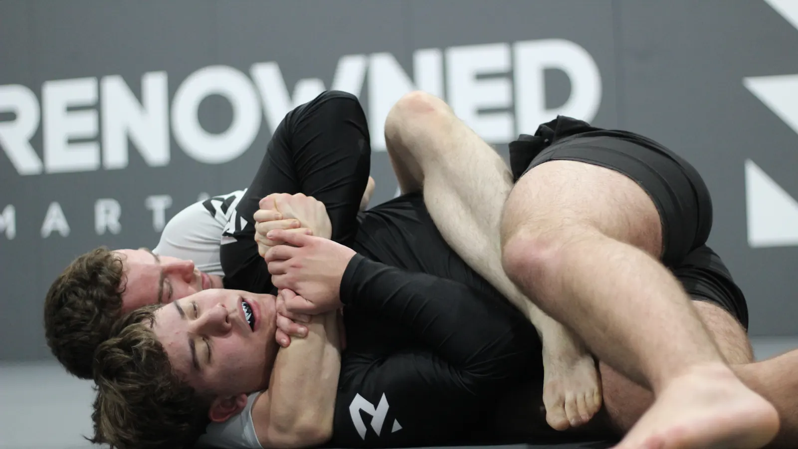 Two adults mid technique on the mats, one working to pass the other's guard